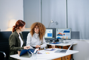 Two businesswomen in modern office analyzing graphs and reports on laptops. Real people collaborating on data, marketing,