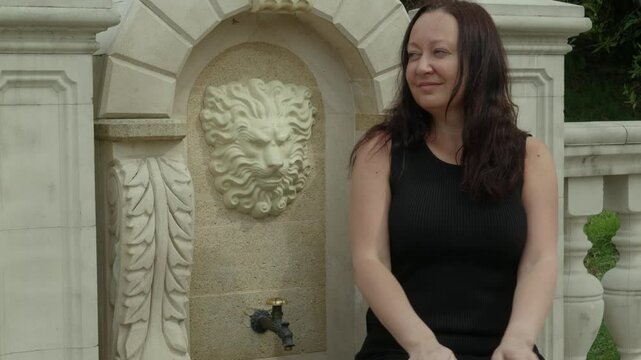 Tourist enjoying a moment of pause in a public park, refreshing herself at a fountain decorated with a lion head bas relief and acanthus leaves, a symbol of strength and prosperity