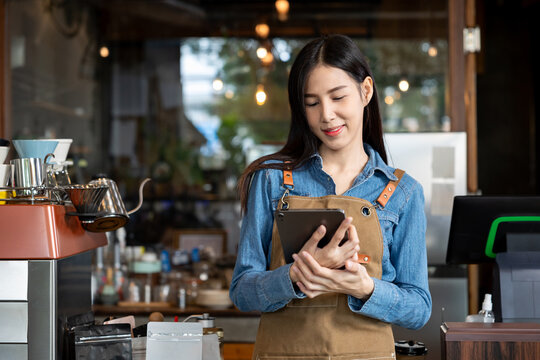 Asian female barista in brown apron using digital tablet while standing behind coffee counter with espresso machine and packaging supplies. Calm mood in cozy cafe during work preparation.