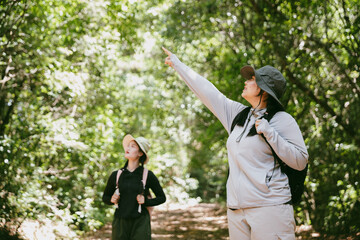 Two female friends with backpacks on vacation hiking through countryside together. Asian friend girls backpacker friend travel in forest wild together. happy and enjoying sunny day while hiking.