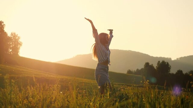 Young beautiful girl tourist spins on a green field at sunset with her arms raised, spins and enjoys the beautiful summer mountain landscapes.