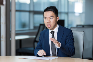A young man in a suit gestures while speaking during a meeting in a modern office. His expression...