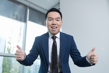 A young man in a suit leans forward with a wide smile, giving two thumbs up in a well-lit office....
