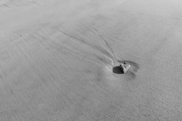 Lone pebble rests on a smooth, rippled sandy beach in black and white, creating subtle lines and textures around it