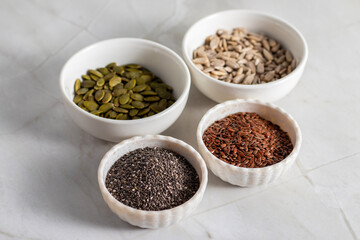 Four white ceramic bowls contain different seeds—pumpkin, sunflower, chia, and flax—arranged neatly on a light marble surface with a minimal aesthetic.
