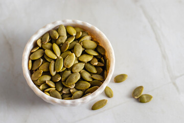 A white ceramic bowl filled with green pumpkin seeds sits on a light marble surface, with a few seeds scattered casually around the bowl.
