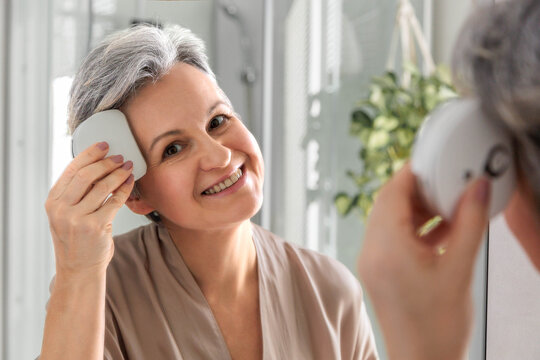 miling mature woman using facial massage device in front of bathroom mirror at home