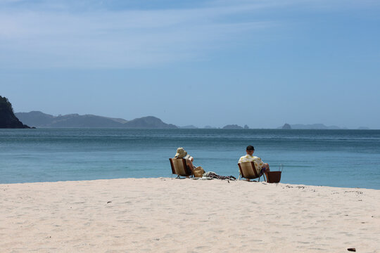 Couple reading on the beach with camping chairs facing the ocean. Relaxing day in Hahei beach, sunny view of the islands and clear blue summer sky. Beautiful Coromandel region in New Zealand.