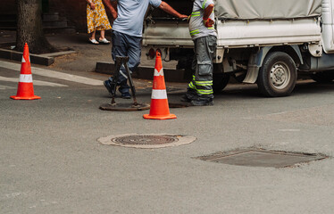 Urban utility workers servicing underground water infrastructure at an open manhole. Safety cones, service van, and tools visible. City maintenance without visible faces or brands