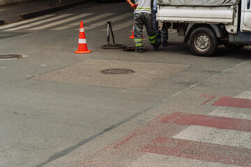 Urban utility workers servicing underground water infrastructure at an open manhole. Safety cones, service van, and tools visible. City maintenance without visible faces or brands