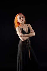 A woman in a black dress with red hair poses for the camera in a dark room. She has a beautiful...