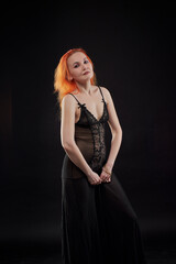 A woman in a black dress with red hair poses for the camera in a dark room. She has a beautiful...