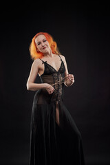 A woman in a black dress with red hair poses for the camera in a dark room. She has a beautiful figure. The girl is not wearing underwear under her dress.