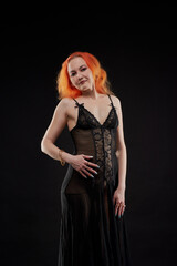A woman in a black dress with red hair poses for the camera in a dark room. She has a beautiful...