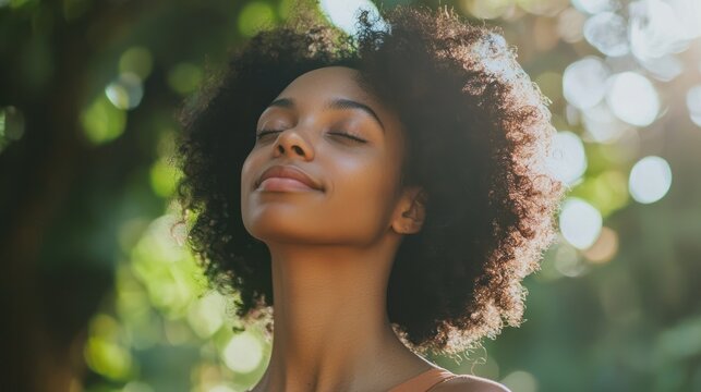 A young woman with curly hair standing outdoors in a lush green park, wearing a brown top, with her eyes closed and a serene expression on her face.