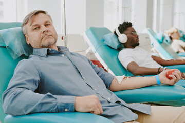 Middle aged Caucasian man donating blood while reclining in medical chair, holding stress ball,...