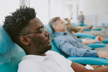 Obraz premium Young Black man donating blood in medical facility, sitting in reclining chair with needle in arm, middle aged Caucasian men donating blood in background, participating in blood donation
