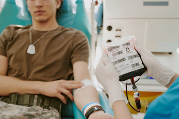 Caucasian teenage boy sitting in medical chair donating blood, while healthcare worker wearing gloves holding blood bag labeled with blood type and barcodes during donation process