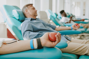 Obraz premium Caucasian middle aged man and Black young adult man donating blood in medical facility, closeup of Caucasian male hand squeezing red heart shaped stress ball during blood donation process