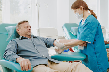 Middle aged Caucasian man donating blood while young female nurse wearing medical mask, assisting...