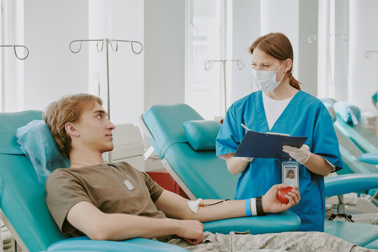Young Caucasian man donating blood while sitting in medical chair, holding red stress ball, female Caucasian nurse in scrubs and mask standing beside him holding clipboard and pen