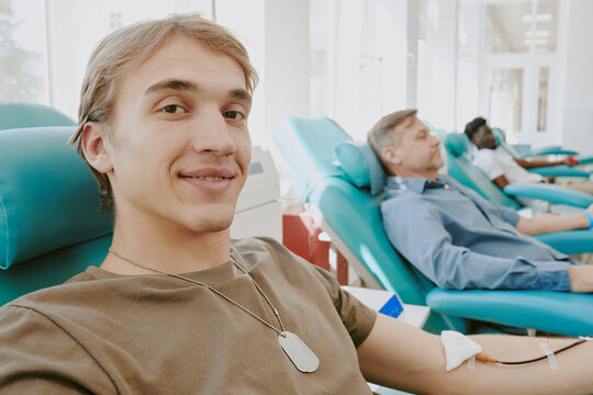 Caucasian young adult man smiling while donating blood in medical clinic, middle aged Caucasian man and Black man sitting in background donating blood, visible medical equipment