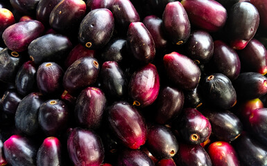 A detailed close-up image of fresh, ripe Java plums, also known as Jamun or black plums (Syzygium cumini), displayed in a pile at a market. The fruits show a natural combination of deep purple, black,