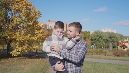 In a bright autumn park, a father gently holds his son, surrounded by colorful foliage and a playground, enjoying quality time on a sunny day.