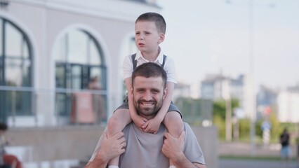 Father and son share joyful moments at a city park, with the child sitting on the father's shoulders, both smiling under the bright sun, surrounded by greenery.