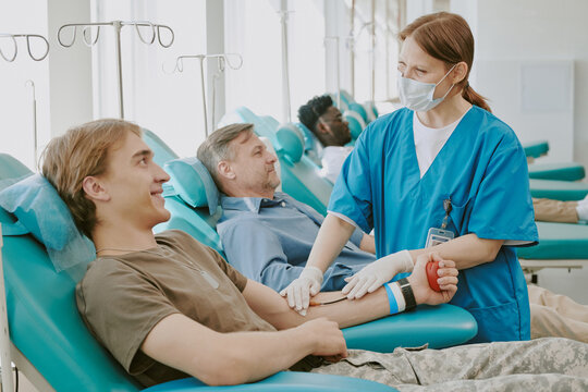 Young Caucasian man donating blood while smiling at nurse, middle aged Caucasian man reclining in background, female nurse wearing medical gloves assisting blood donors in clinic