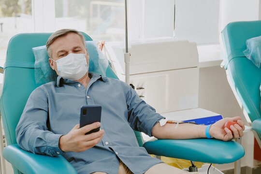 Middle aged Caucasian man donating blood while sitting in medical chair, wearing protective face mask, holding smartphone in hand, receiving blood transfusion through intravenous line