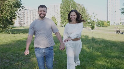 Two people happily stroll together in a park, holding hands and smiling as they enjoy a sunny day surrounded by nature and contemporary architecture.