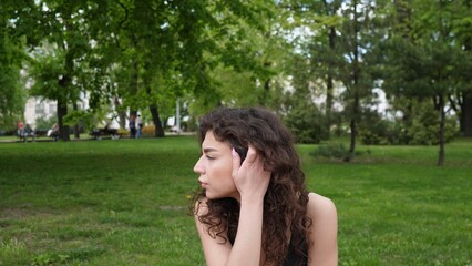 A young woman with curly hair sits alone in a park, surrounded by vibrant greenery and trees, lost in thought as she connects with nature.
