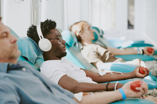 Multiethnic group of young adult and middle aged men donating blood while sitting in medical chairs, holding red stress balls, Black man wearing headphones relaxing during blood donation