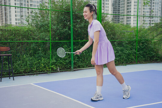 Young woman practicing badminton on court surrounded by lush greenery in an urban setting during a bright day