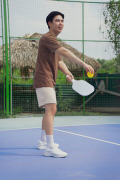 Young Asian man enjoys pickleball on a sunny day at a vibrant outdoor court surrounded by greenery