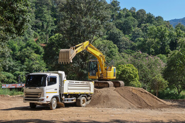 A white truck is waiting for a yellow backhoe to load sand into the back of a construction site surrounded by forest.