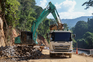 A white truck is waiting for a green backhoe to load sand into the back of a construction site surrounded by forest.