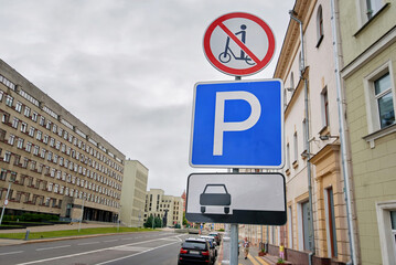 Pole in downtown street displays signage for traffic and pedestrian rules including no scooter zone and specific parking regulations. Pole with regulatory signs, no scooter riding, parking lot symbols