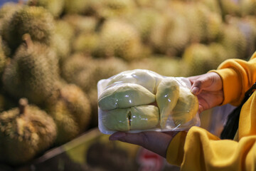  A yellow peeled durian wrapped in clear plastic is in the customer's hand. Background: Many durians in the market in Chiang Mai, Thailand. Thai fruit gardens are sold in a night market