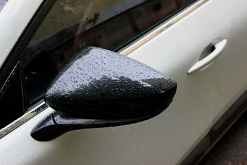 The black side mirror of a white car is covered with large raindrops