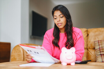 Woman planning finances at home while reviewing bills with a pink piggy bank on the table in the...
