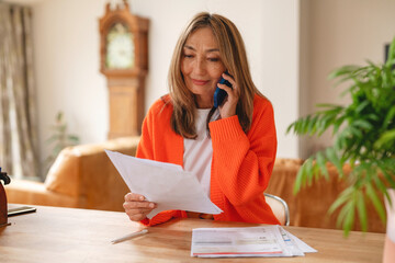 Woman in orange sweater making a phone call while reviewing important documents at a cozy home...