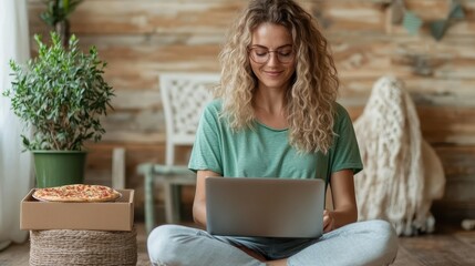 A cheerful young woman sits on the floor, happily working on her laptop with a pizza nearby, embodying comfort and productivity in a cozy setting.