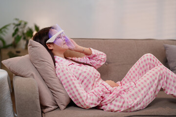Fototapeta premium Asian adult woman enjoying relaxing LED light therapy mask on sofa for enhanced longevity skincare routine at home