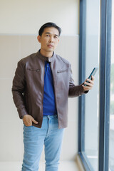 Fototapeta premium Confident adult Asian businessman wearing a stylish brown leather jacket, holding a smartphone while standing next to a window in a modern office building during the day