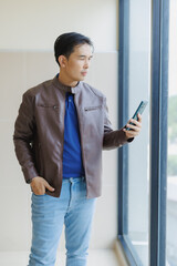 Fototapeta premium Young Asian businessman dressed in casual attire, using his mobile phone while standing beside a window in a modern office building, enjoying natural light and a bright atmosphere
