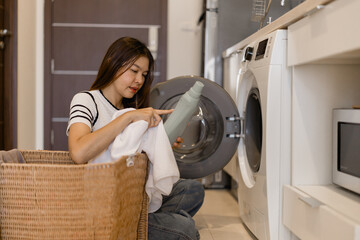 Young woman sitting on floor next to washing machine and laundry basket, holding white towel and...
