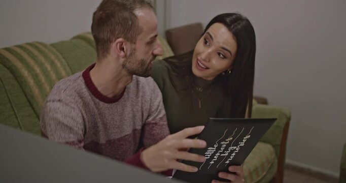 A young couple sits on a sofa at home, reminiscing over sweet memories in a photo album.