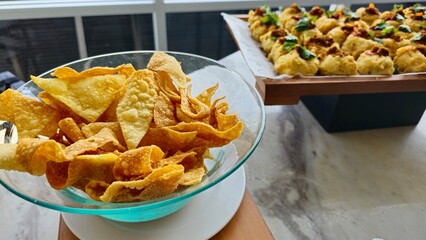 Crispy wonton chips in a glass bowl with assorted savory appetizers in the background, perfect for buffet, catering, or Asian-inspired food presentations.